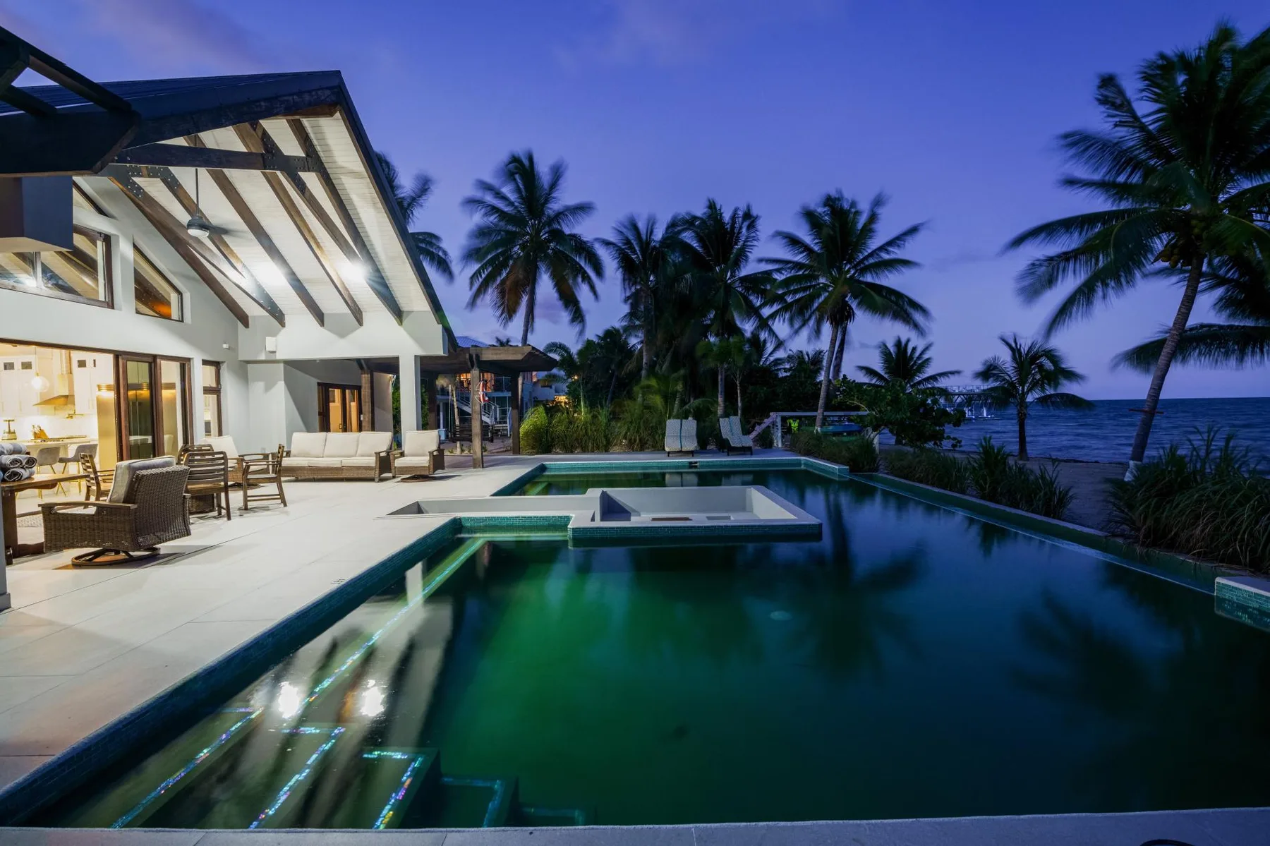 A modern house opens onto a swimming pool, with palm trees and the sea beyond.