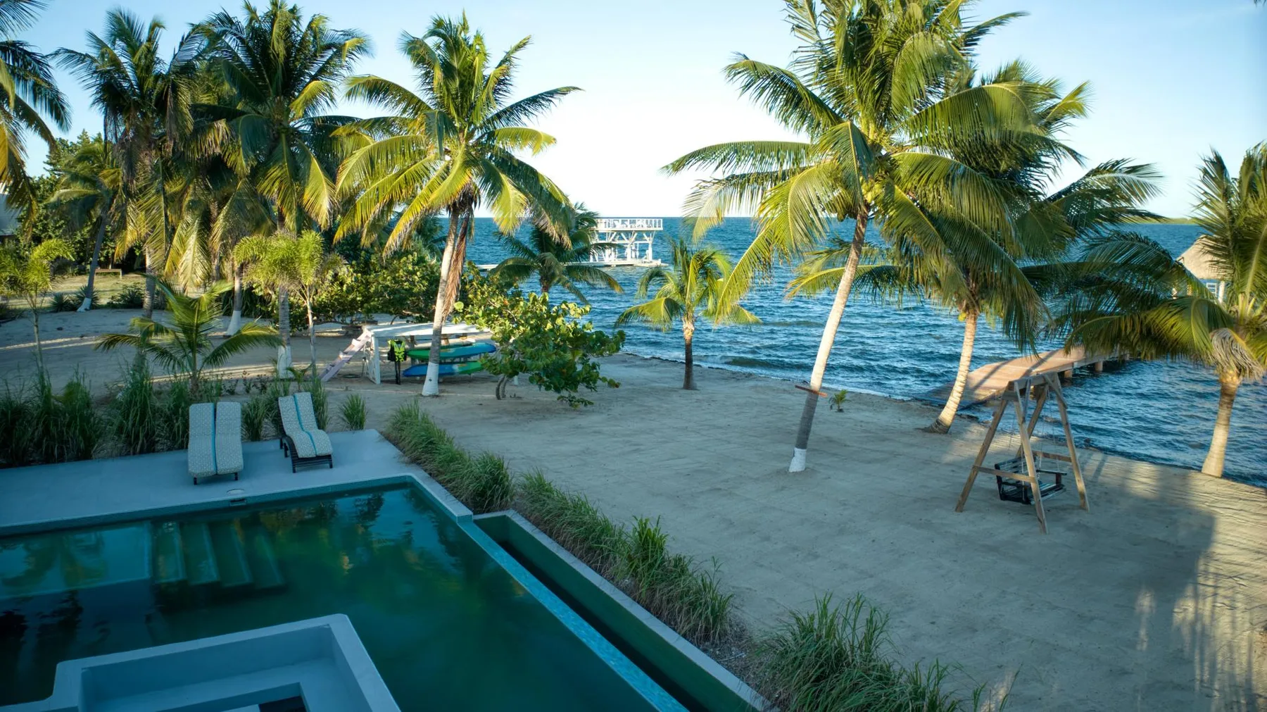 A sandy beach with palm trees overlooks blue water beside a pool.