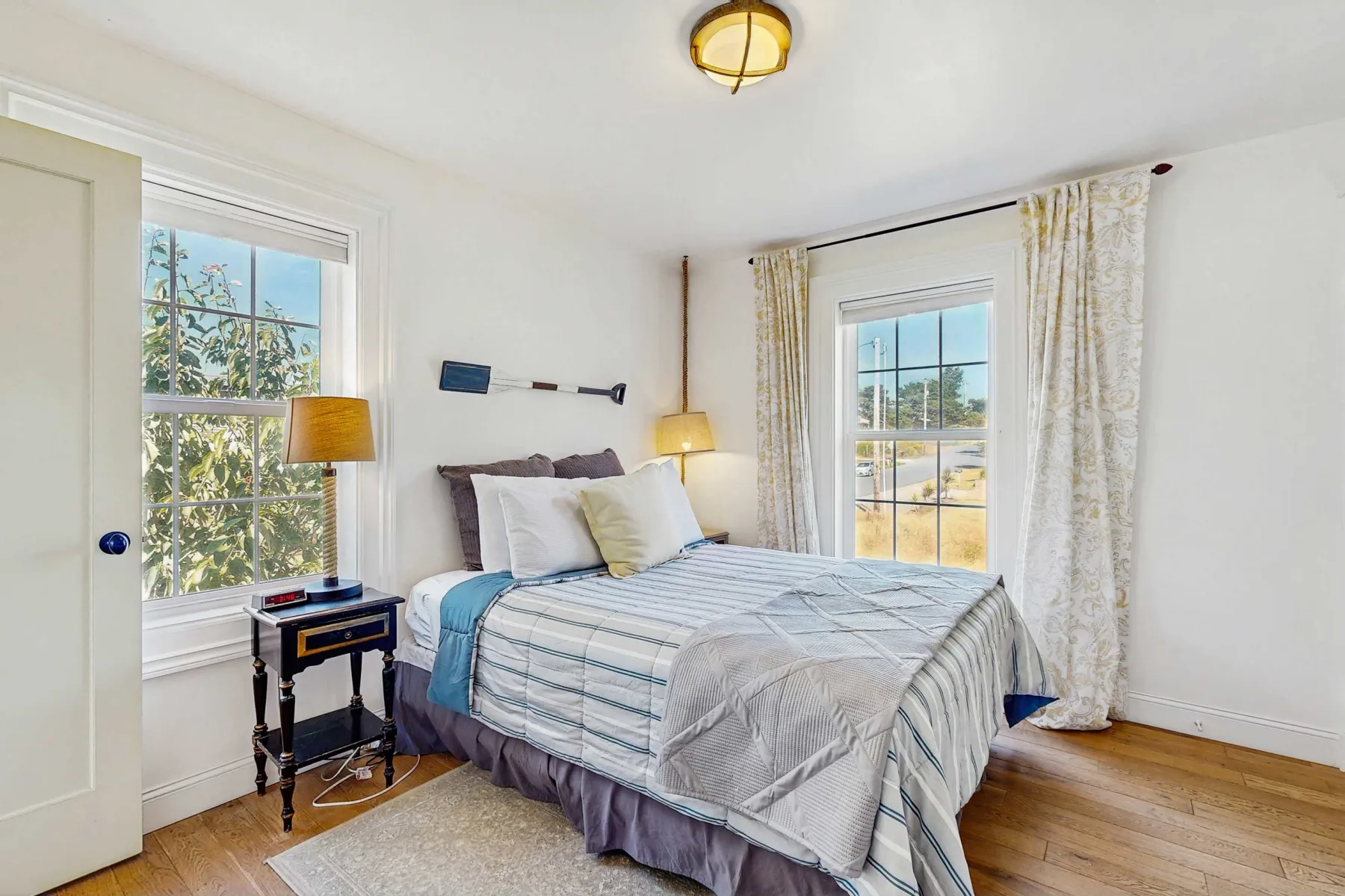 Un dormitorio con una cama bien tendida, mesitas de noche y ventanas con vista a árboles.