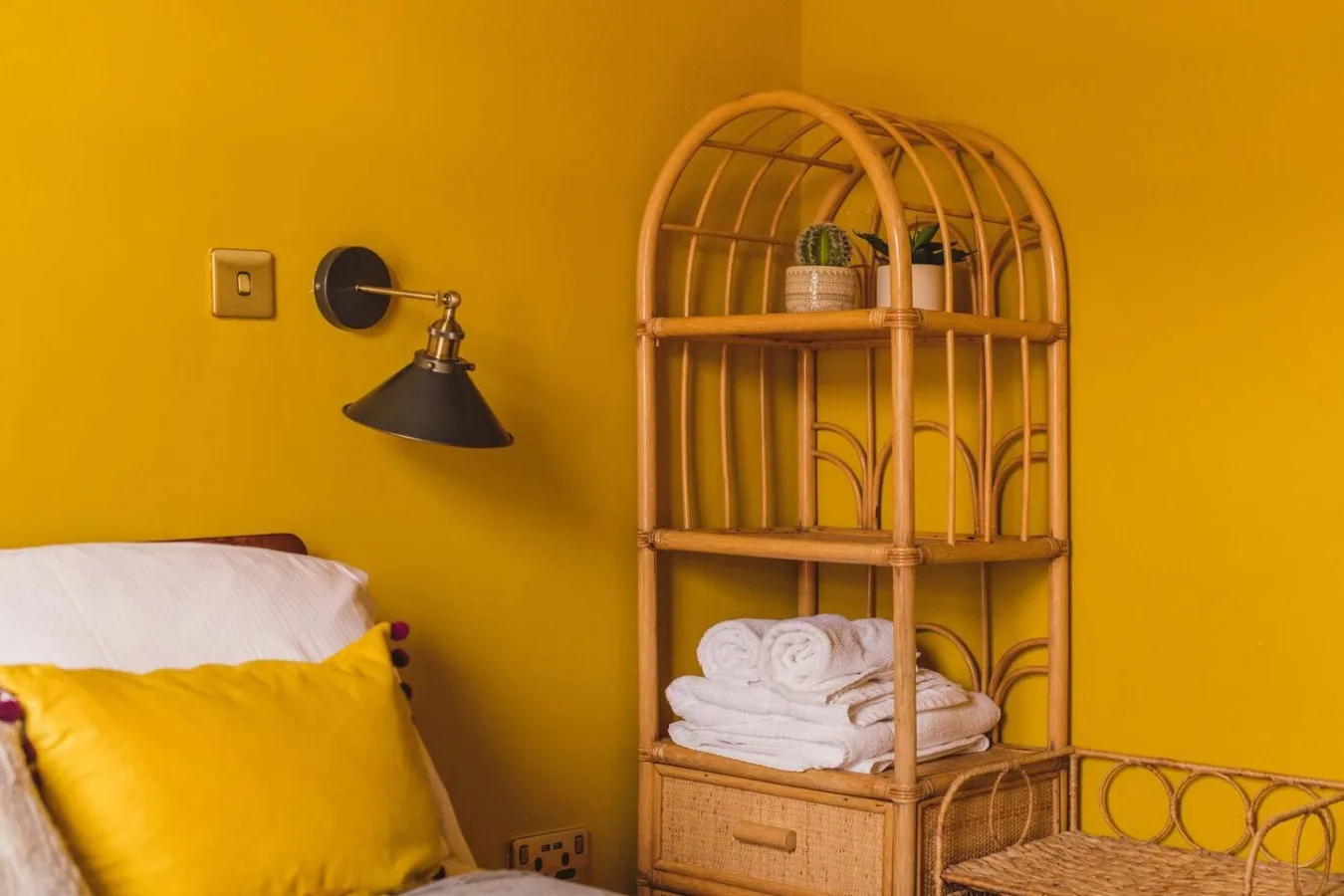 A bedroom corner with a yellow wall, bedside lamp, and woven shelving.