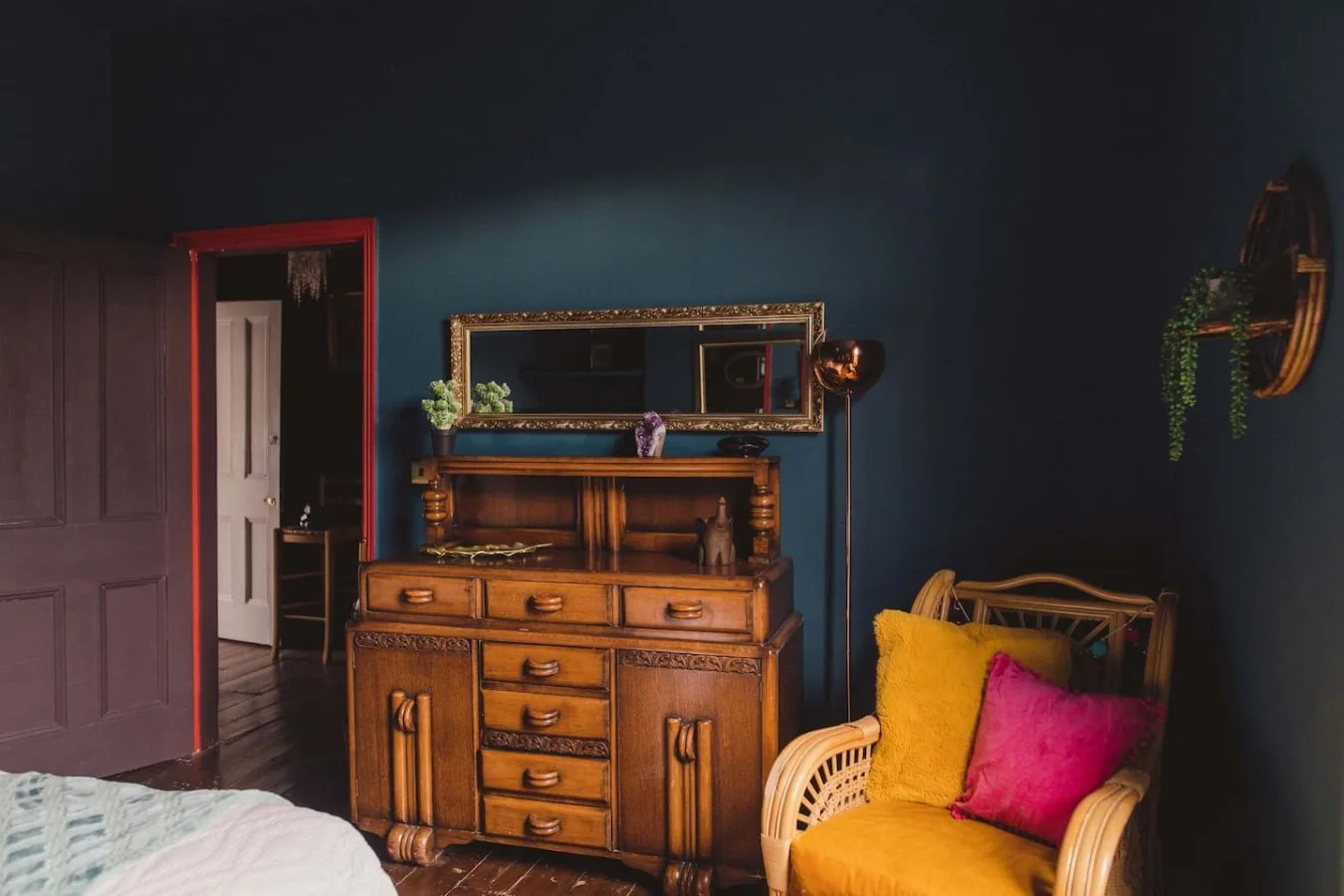 A dark blue bedroom features a wooden dresser and a yellow chair with colorful cushions.