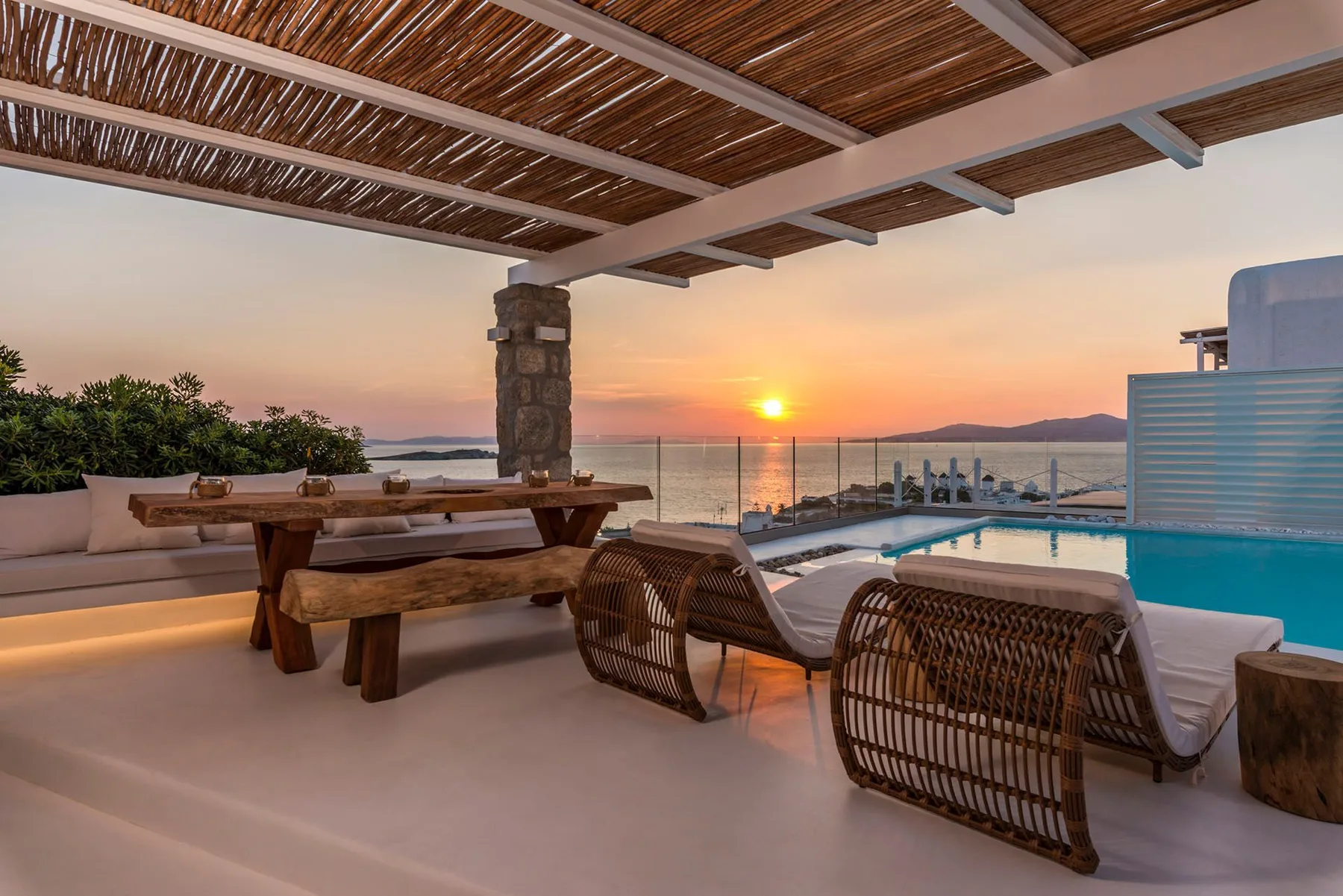 A covered outdoor terrace overlooks a pool and the sea at sunset.