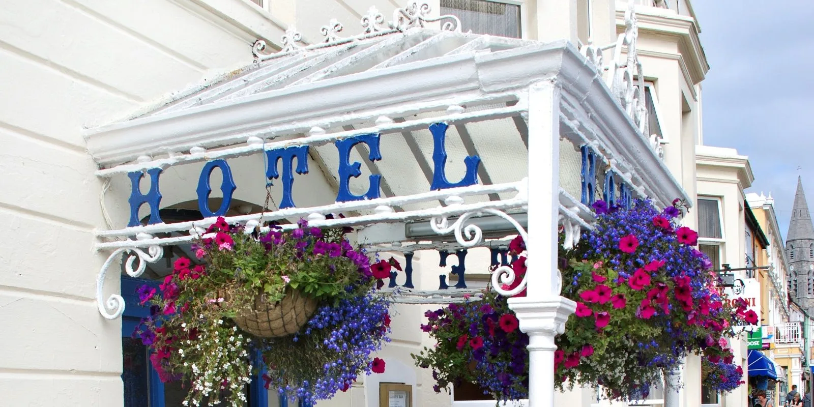 Espacio representativo del hotel: Entrada de hotel con adornos blancos ornamentados y coloridas cestas de flores colgantes.