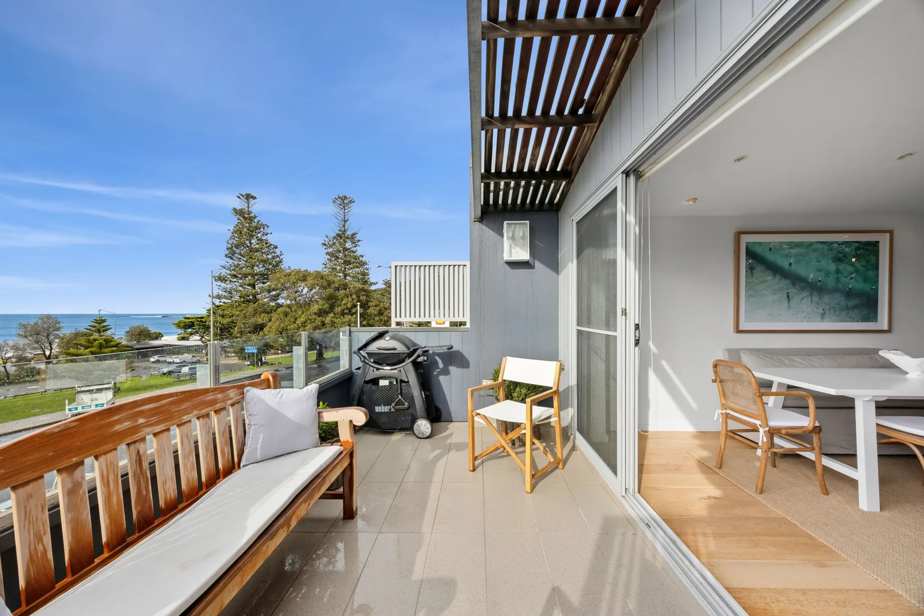 A balcony with outdoor seating overlooks trees and the ocean, beside an open dining area.