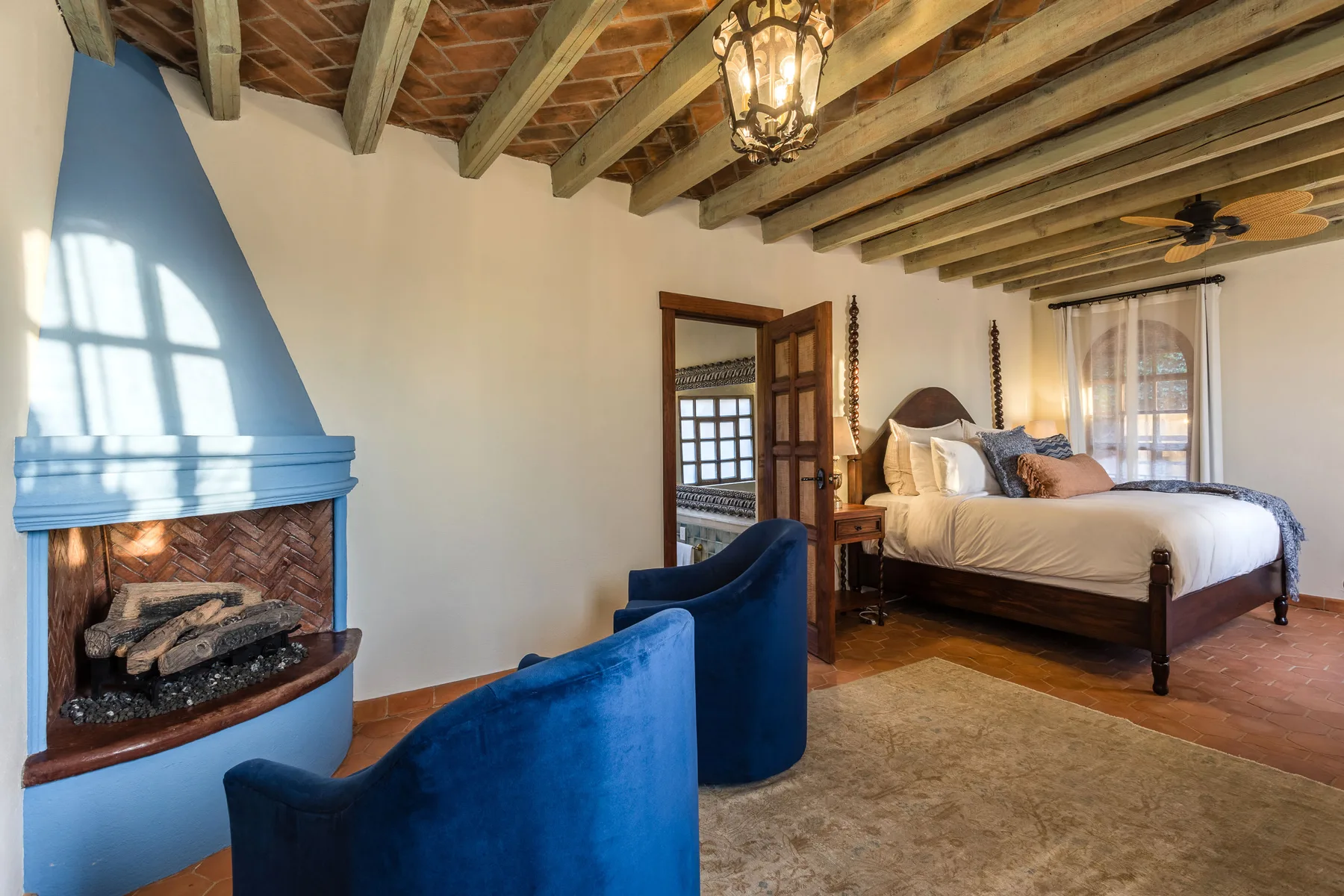 A bedroom with a bed, blue seating, and a fireplace beneath exposed ceiling beams.