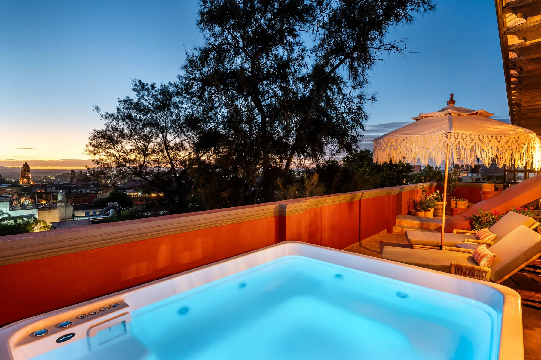A rooftop terrace features a lit hot tub beside lounge seating and a fringed umbrella.