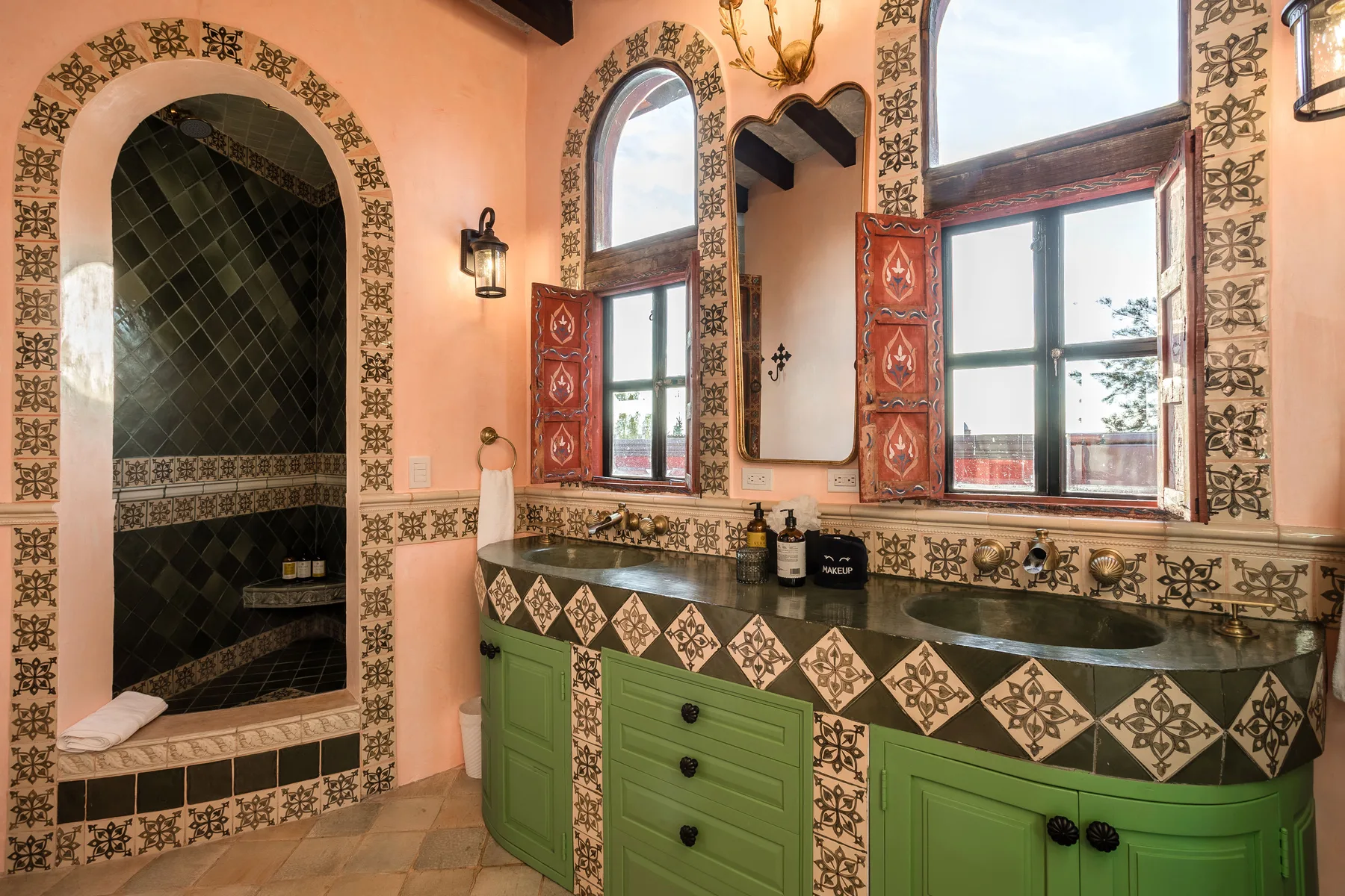 A tiled bathroom vanity with green cabinetry sits beneath large windows.