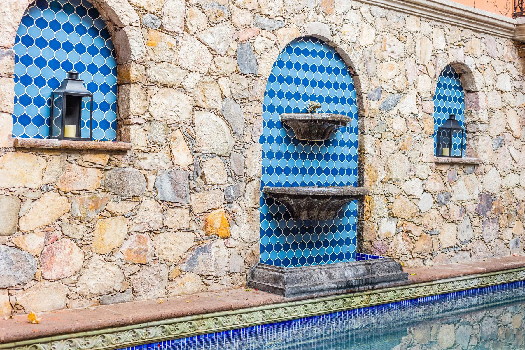 A stone wall with blue-tiled arched niches and a fountain beside a pool.