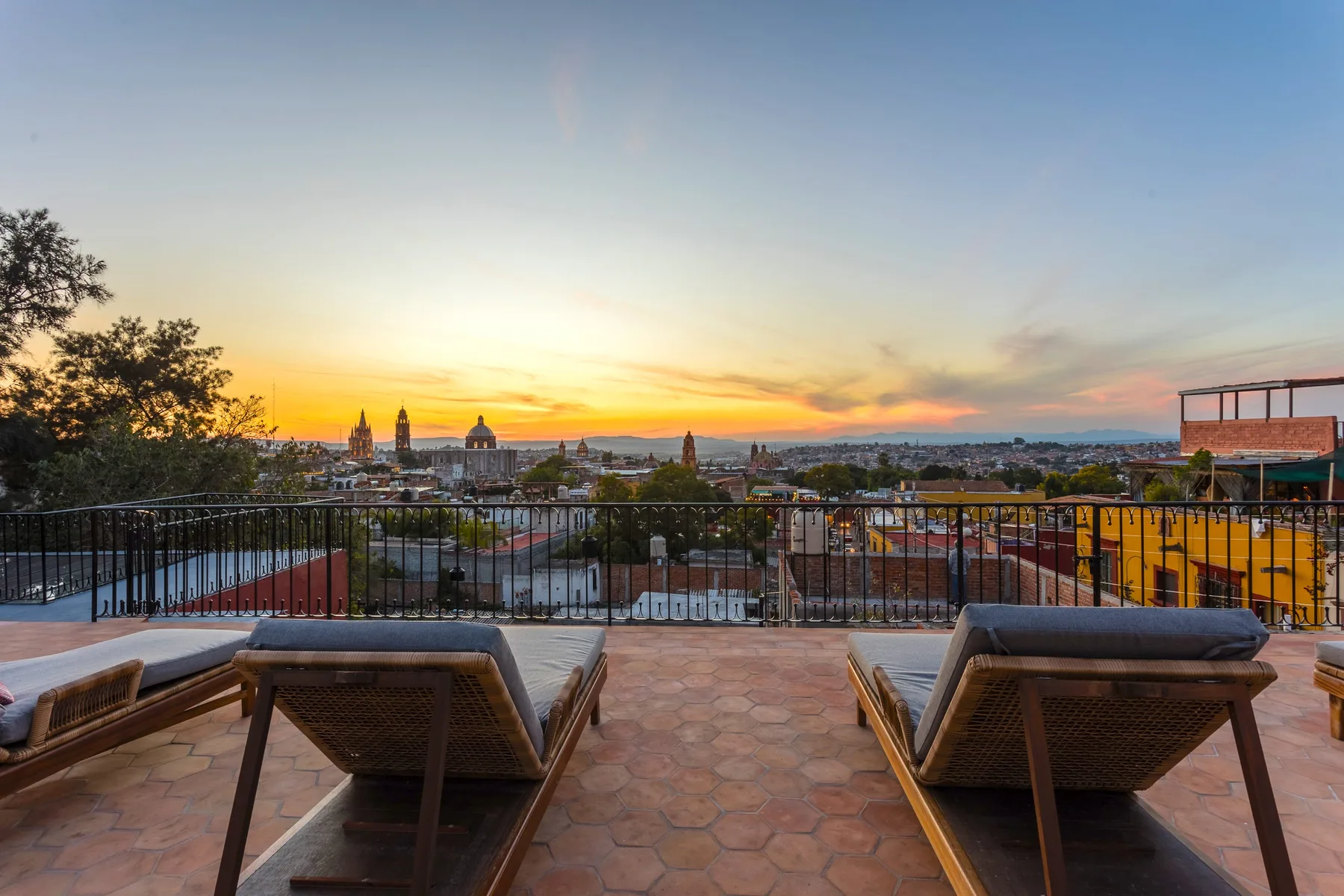 A rooftop terrace overlooks a city at sunset, with lounge seating by the railing.