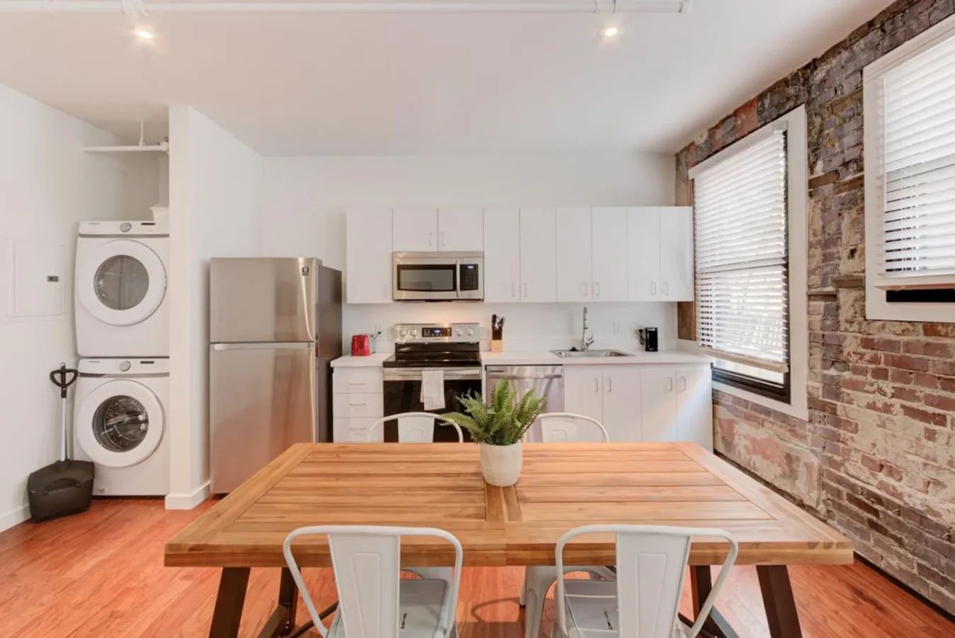 A dining table sits in a bright kitchen with laundry appliances and exposed brick.
