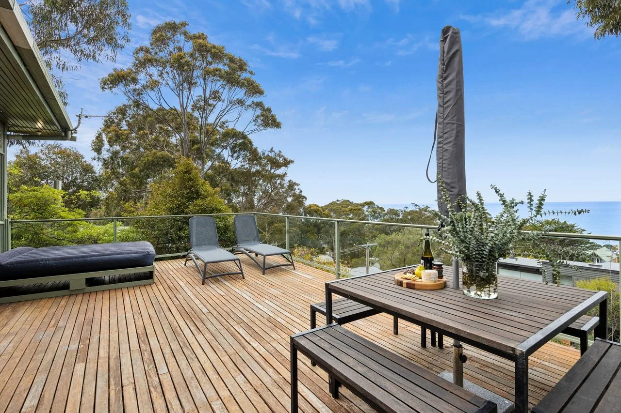 Una terraza exterior con asientos y una mesa da a árboles frondosos bajo un cielo azul.