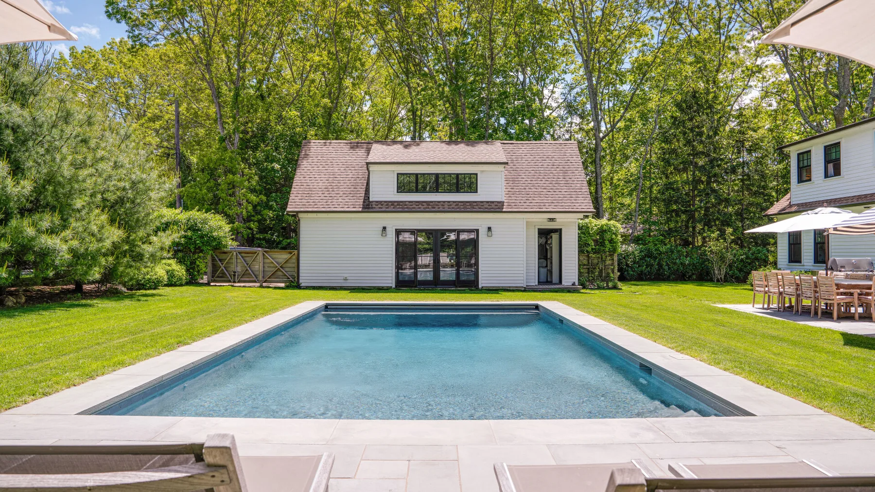 Una piscina en el patio está frente a una casa blanca, rodeada de césped verde y árboles.
