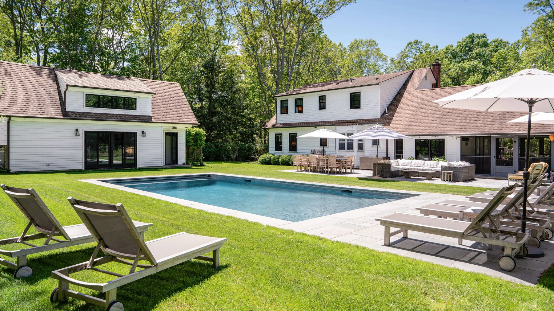 Una zona de piscina al aire libre junto a una casa grande, con tumbonas y asientos de patio.
