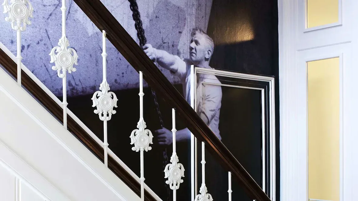 Espacio representativo del hotel: Una escalera con barandillas blancas ornamentadas pasa junto a una gran imagen mural en blanco y negro.