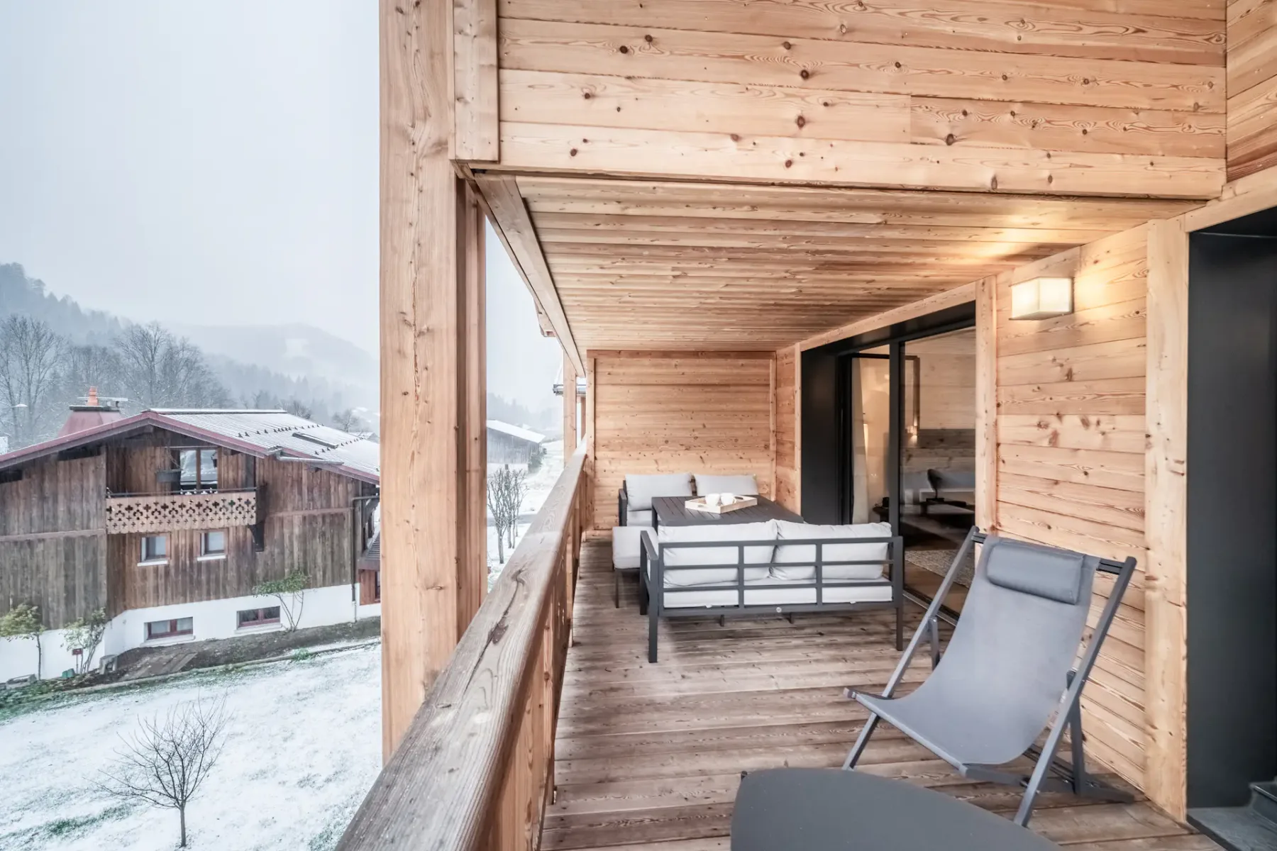A covered wooden balcony with outdoor seating overlooks a snowy landscape.