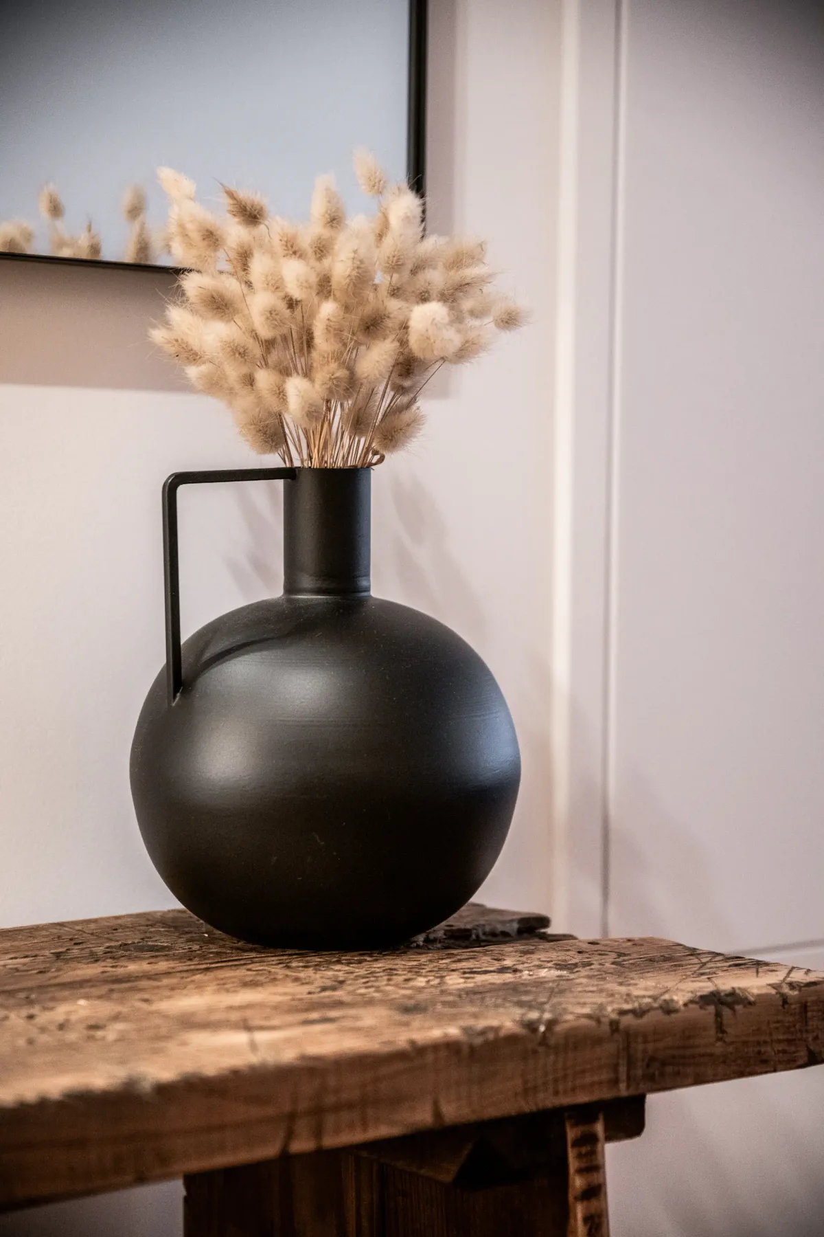 A dark decorative vase with dried flowers sits on a wooden table beneath framed artwork.