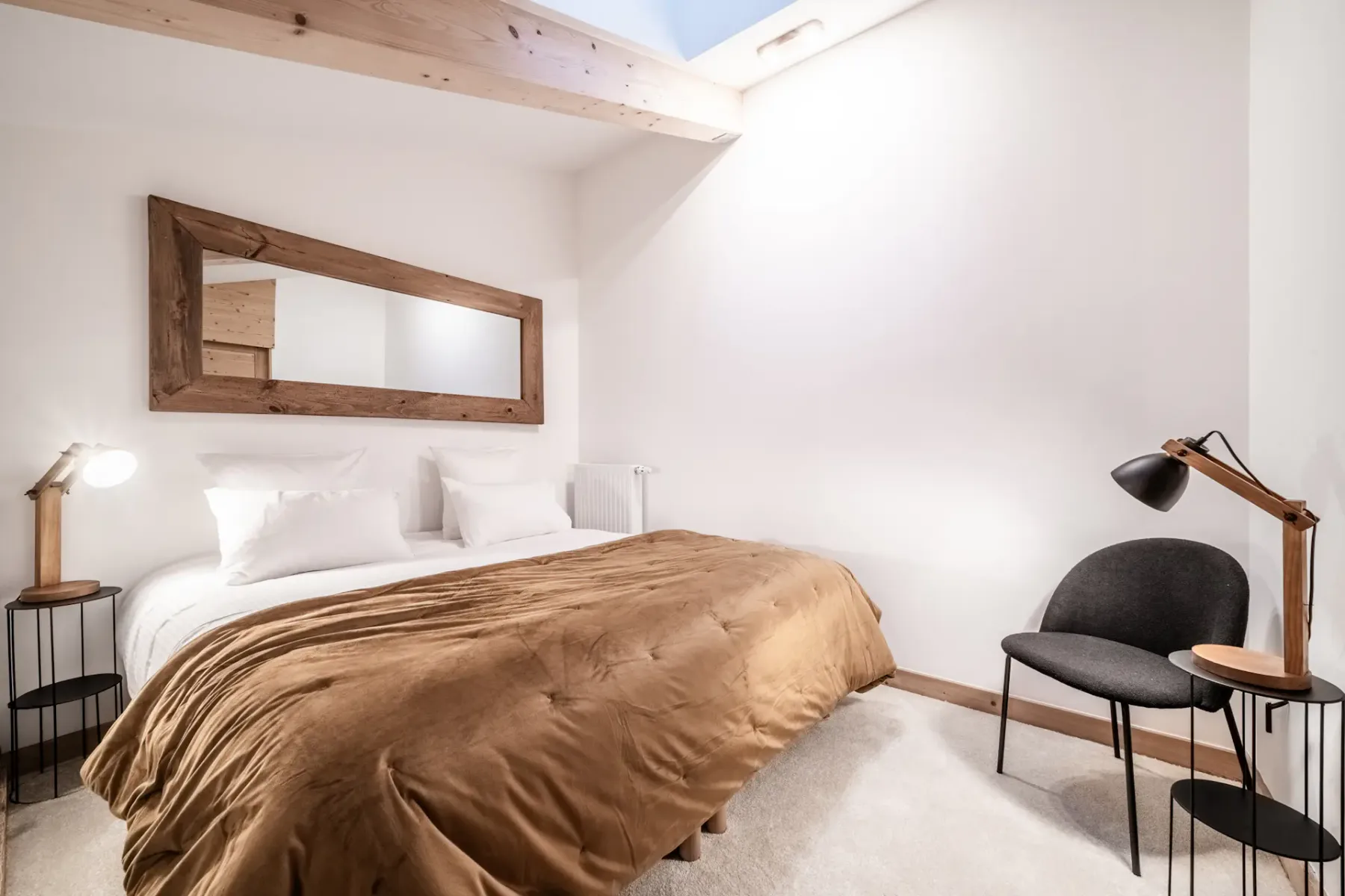 A bedroom with a bed, a wall mirror, and a chair beside a lamp.