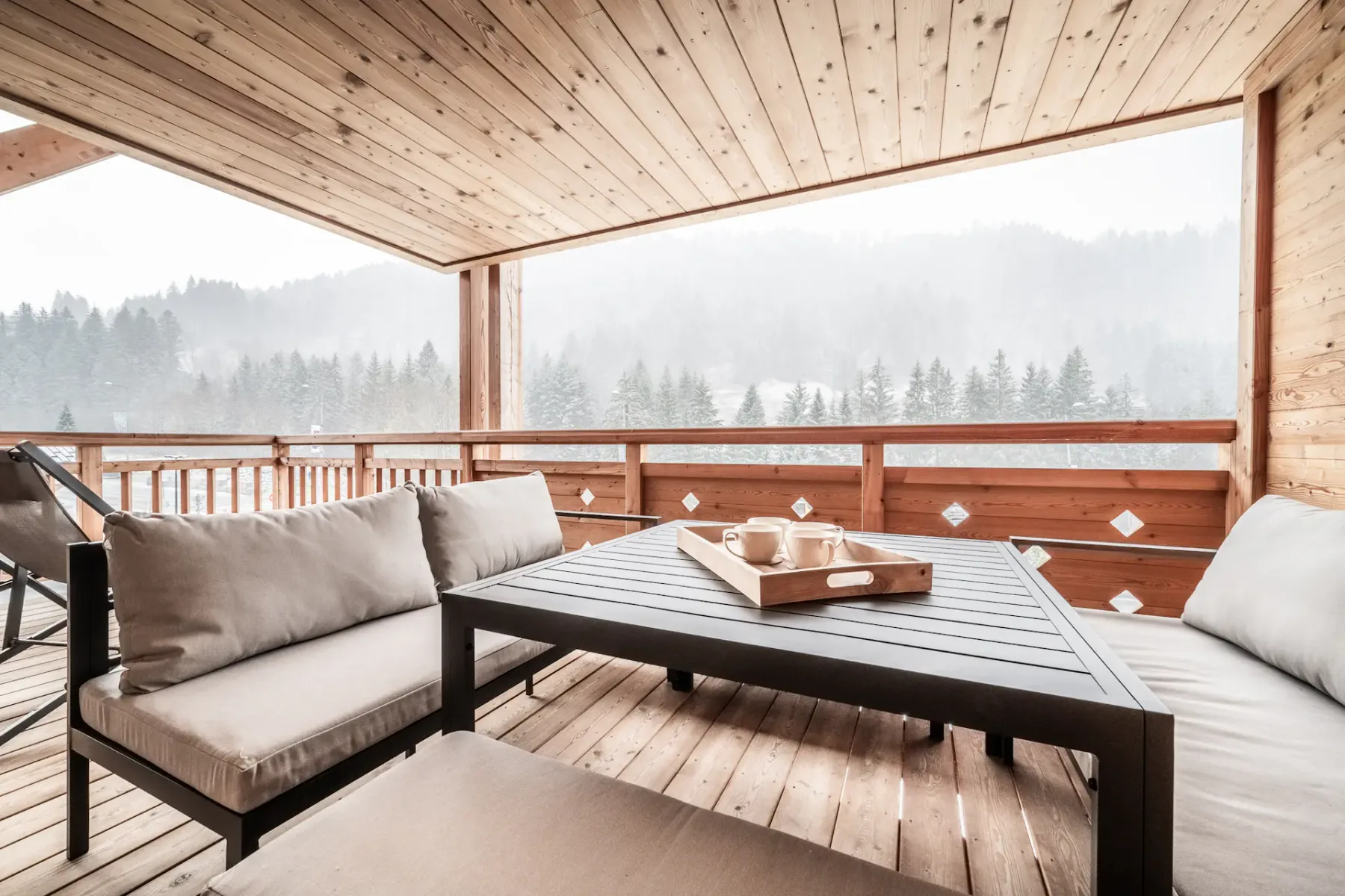 A covered wooden deck has cushioned seating and a low table overlooking a misty forest.