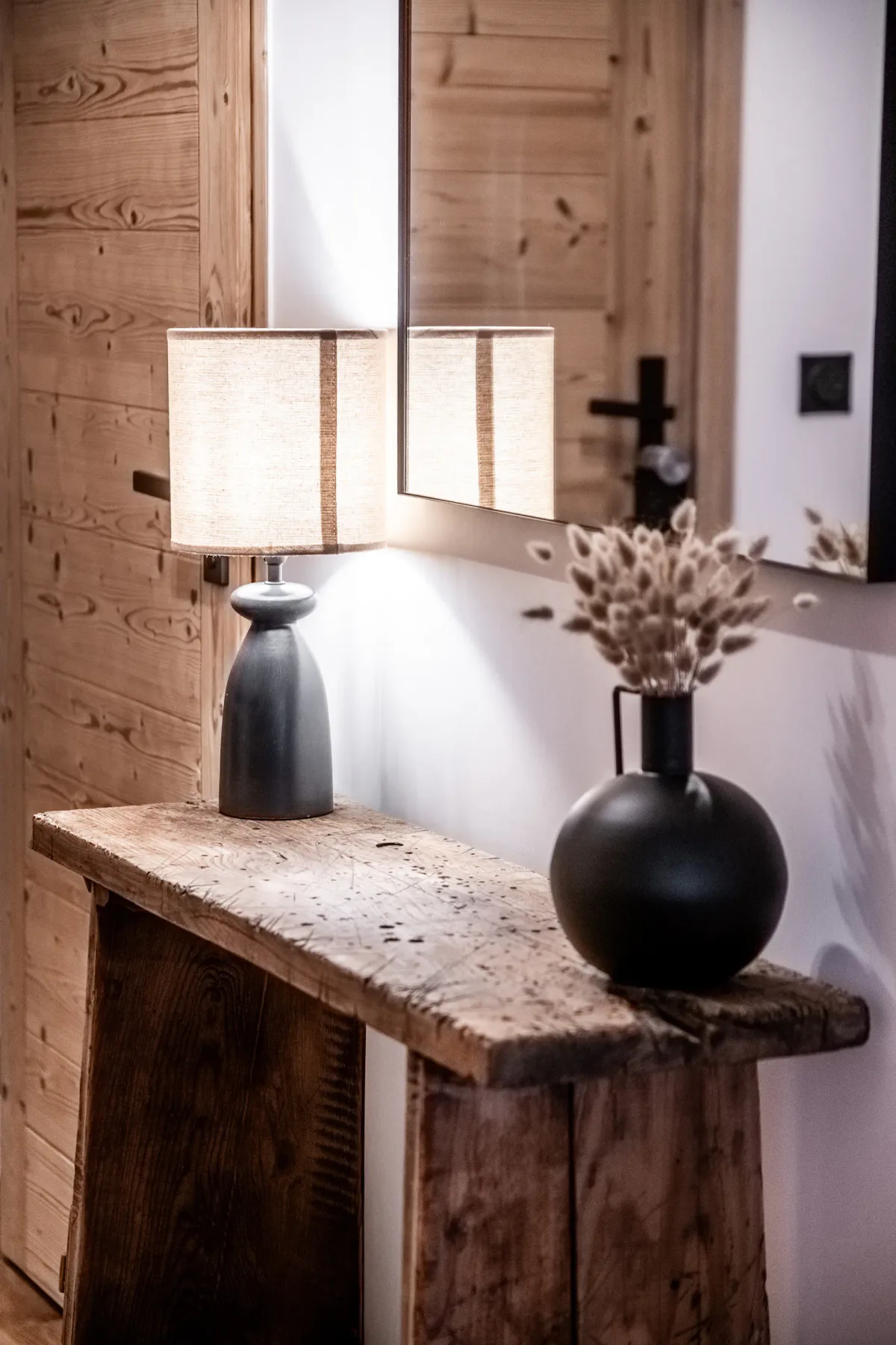 A rustic console table with a lit lamp and a vase of dried flowers.