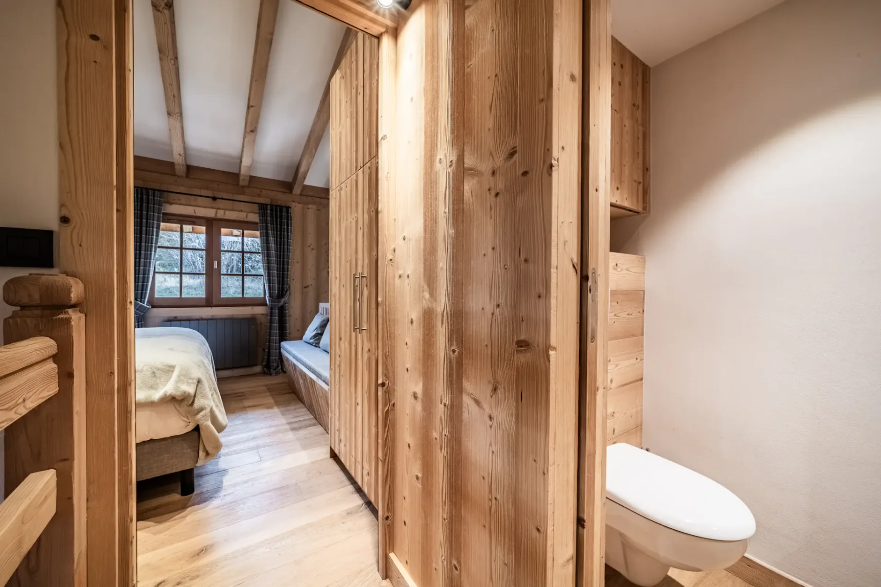A rustic bedroom with a bed, a window, and a toilet visible beyond a wooden partition.