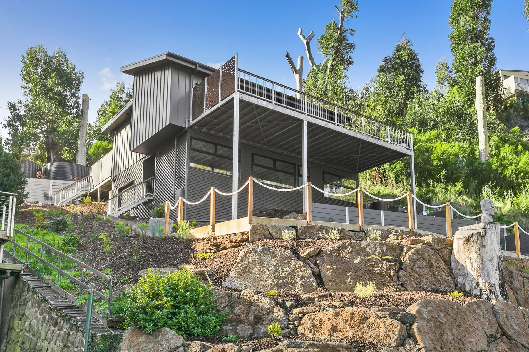 Una casa elevada se alza sobre una ladera rocosa ajardinada y rodeada de árboles.