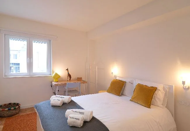 A bedroom with a neatly made bed and a window beside a small desk.