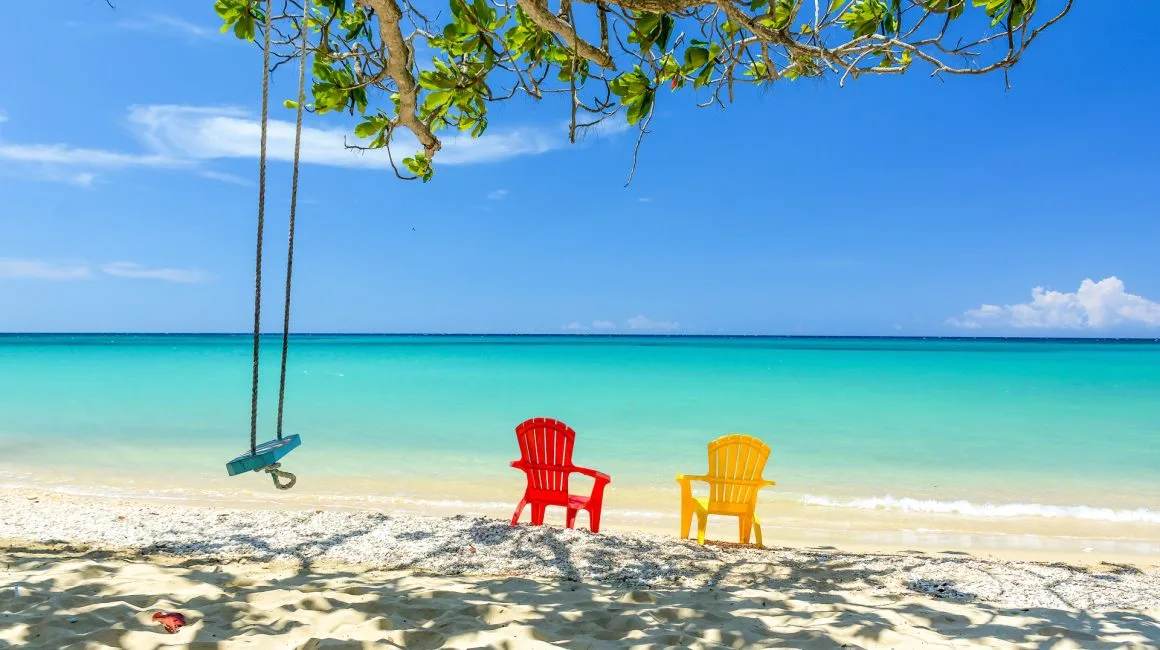 Espacio representativo del hotel: Una playa tropical con dos sillas de colores y un columpio bajo un árbol.