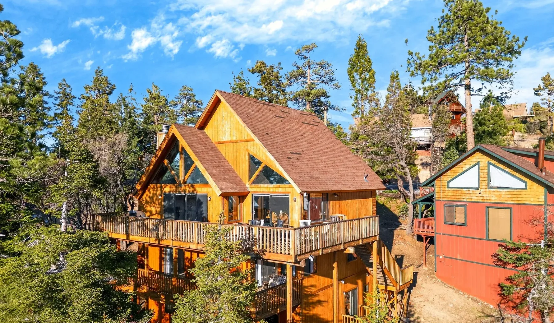 A wooden cabin with a steep roof and wraparound deck sits among pine trees.