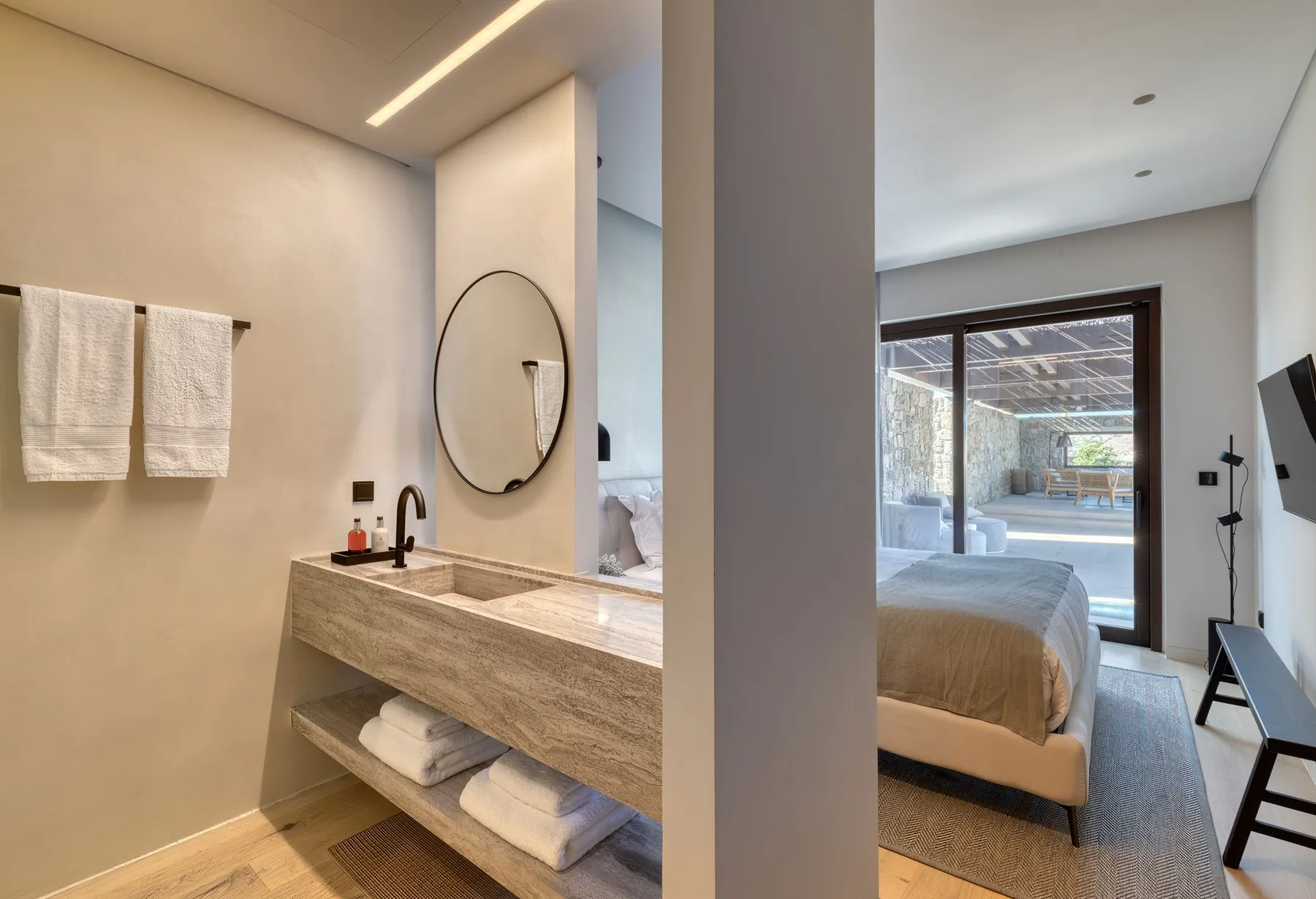 A bathroom vanity with a round mirror opens onto a bedroom with a large window.