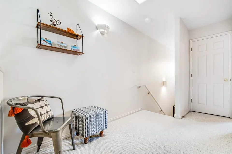 A bright carpeted room has a metal chair and a small upholstered ottoman.