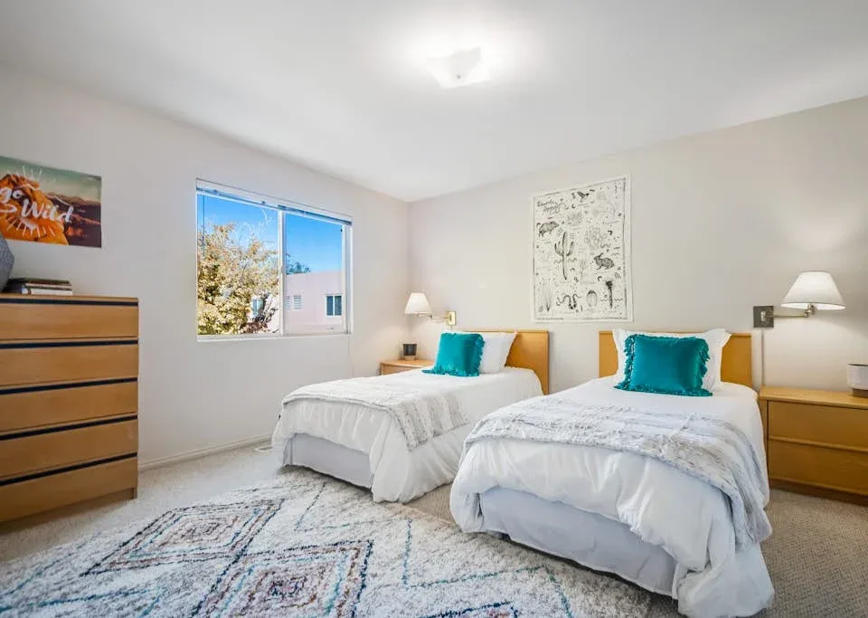 A bright bedroom with two beds, teal pillows, and a window.