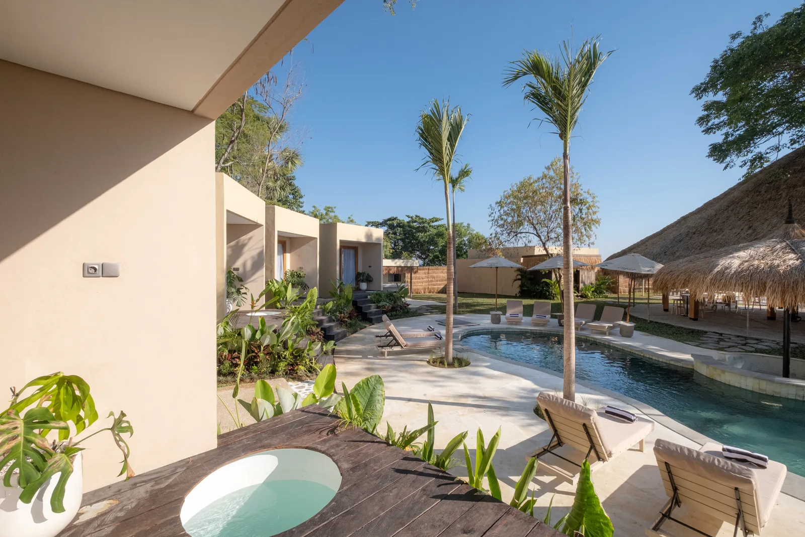 Zona de piscina con palmeras, asientos a la sombra y un pequeño jacuzzi redondo.
