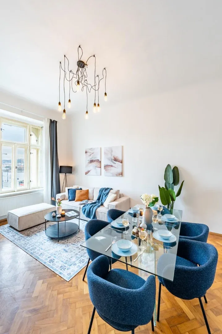 A bright living and dining area features a glass table, blue chairs, a pale sofa, and a large window.