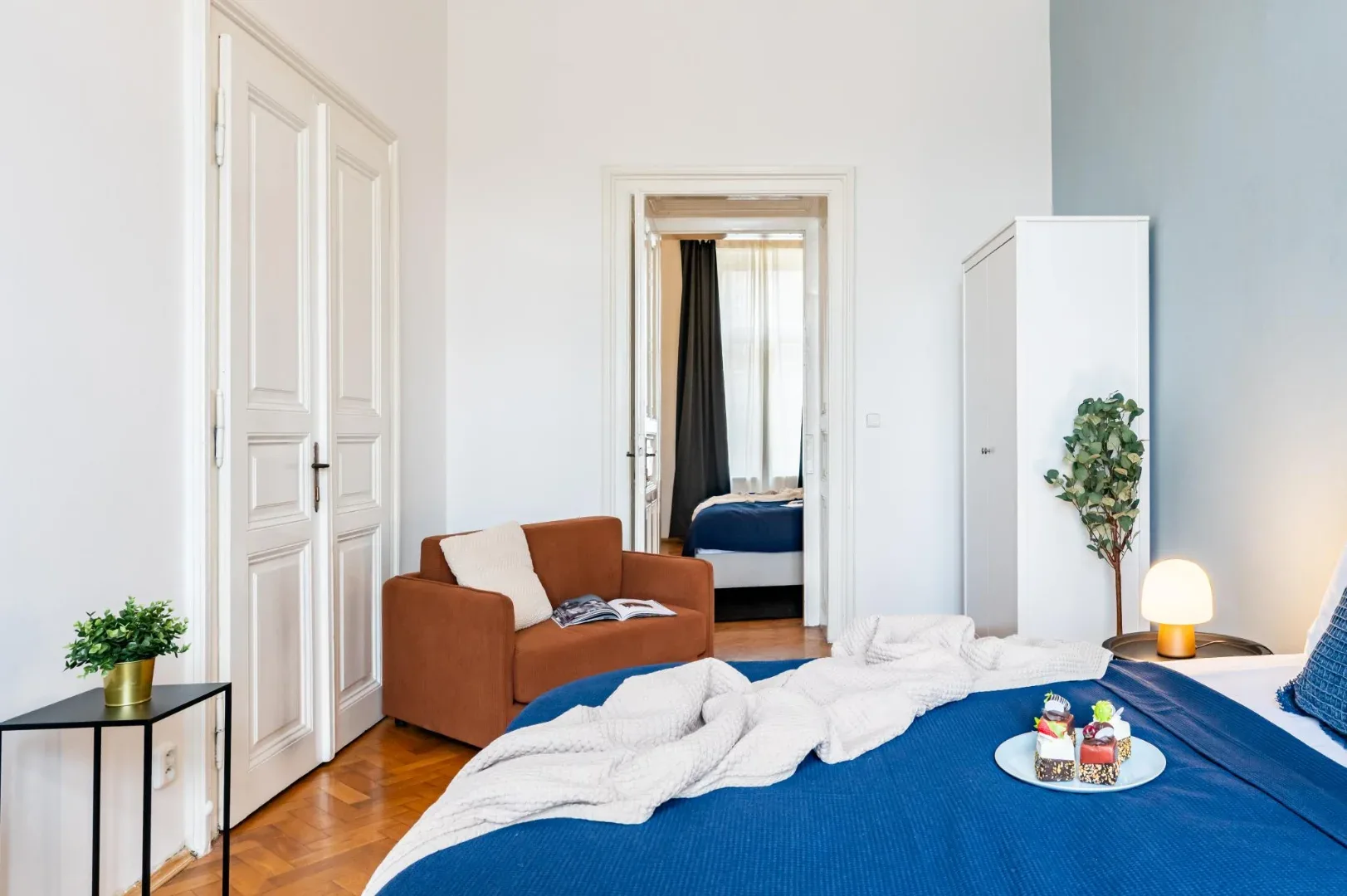A bright bedroom has a blue bed, a rust-colored sofa, white paneled doors, and a wardrobe.