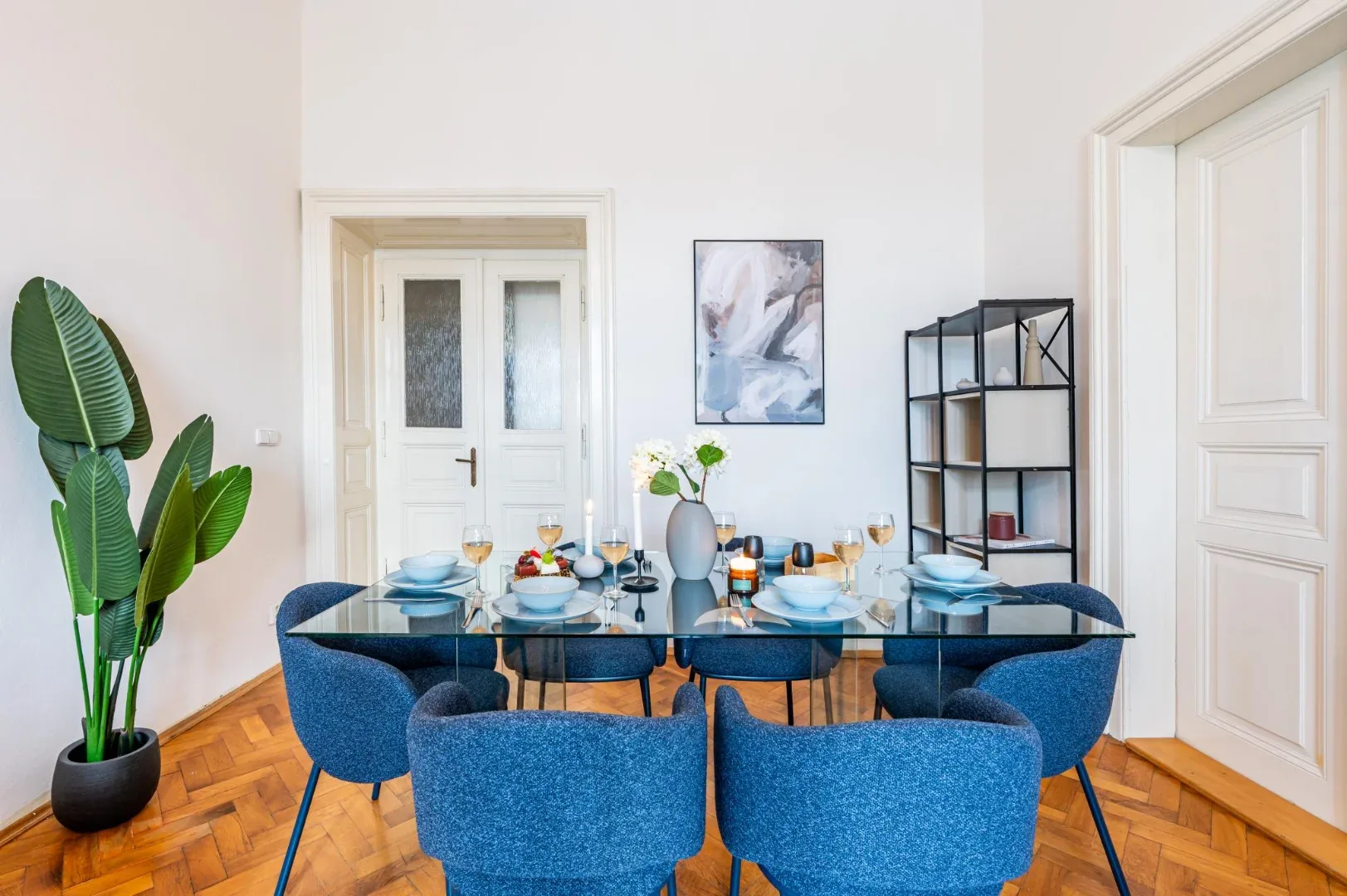 A dining room with a set table, blue upholstered chairs, a potted plant, and an open shelving unit.