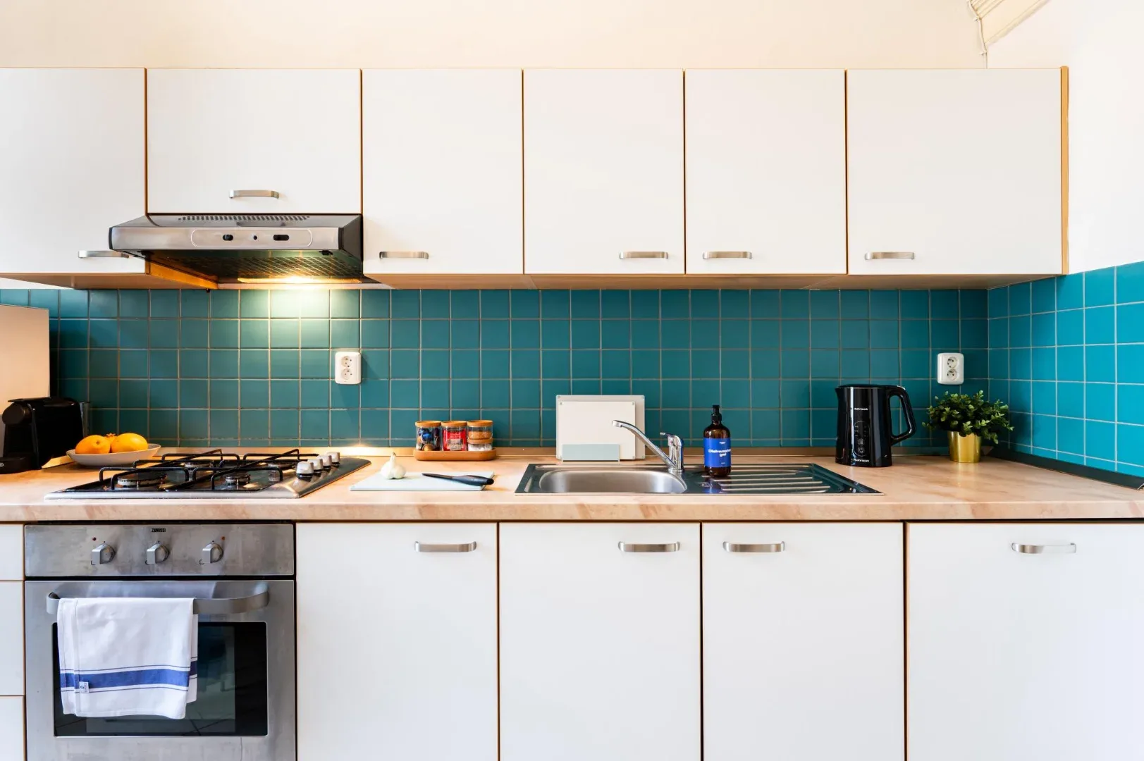 A bright kitchen has white cabinets, a teal tiled backsplash, and a stainless-steel oven and cooktop.