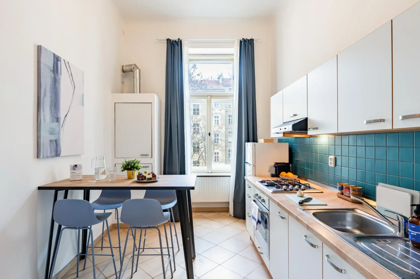 A bright galley kitchen has white cabinets, a blue tile backsplash, a dining table, and a tall window.