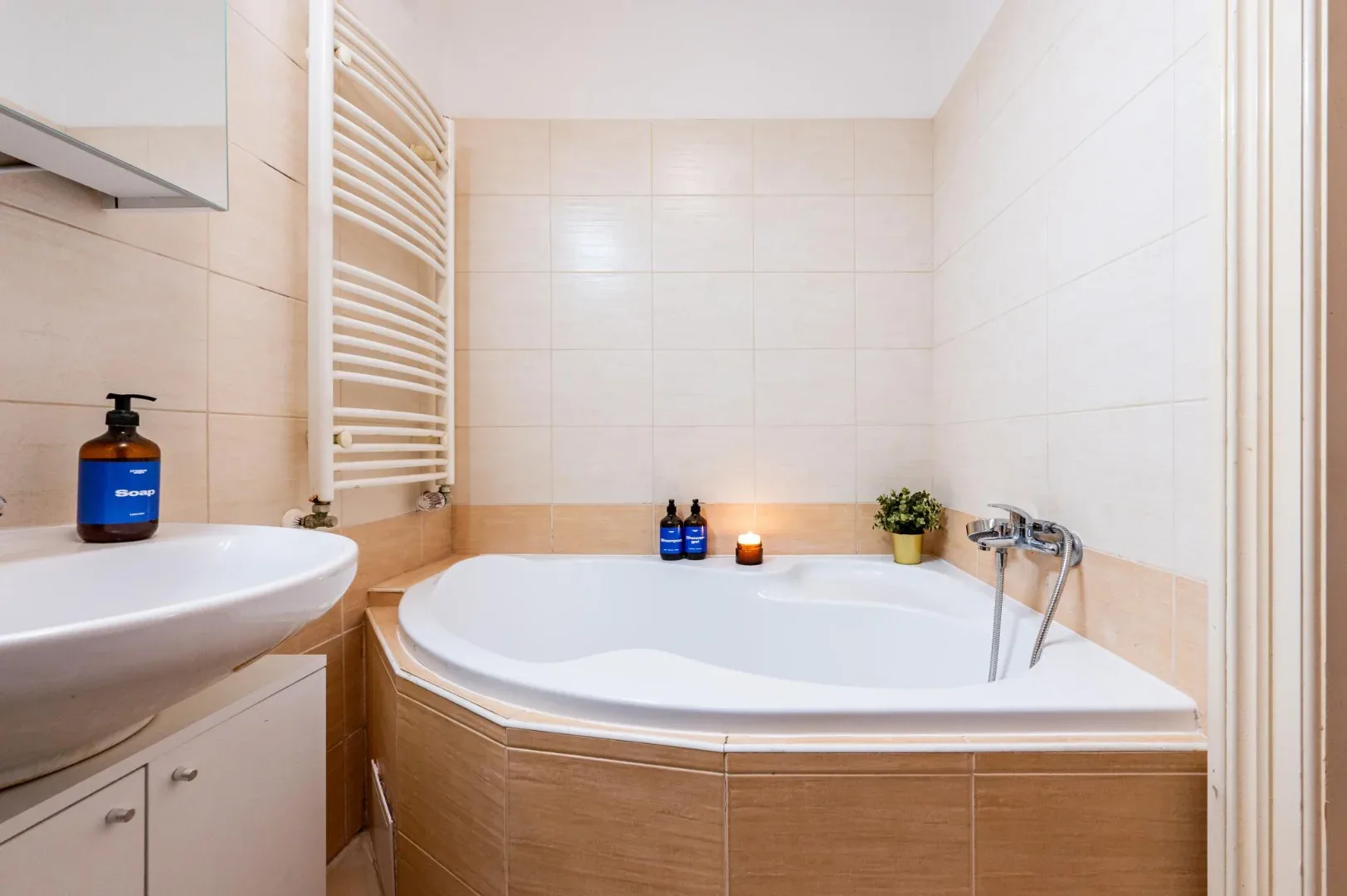 A tiled bathroom features a corner bathtub, wall-mounted towel warmer, and sink.