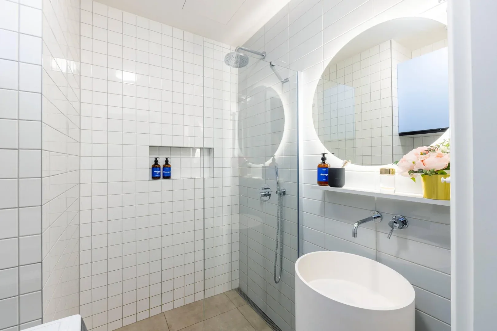 A tiled bathroom has a glass shower, a round mirror, and a white vessel sink.
