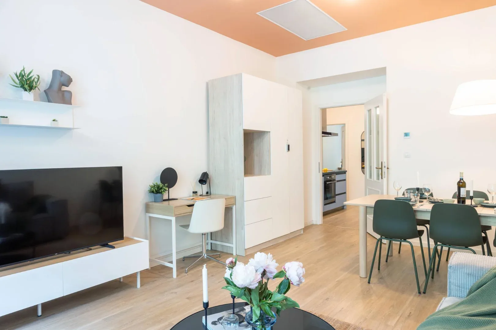 A bright open-plan living and dining area has a television, desk, dining table, and doorway to the kitchen.