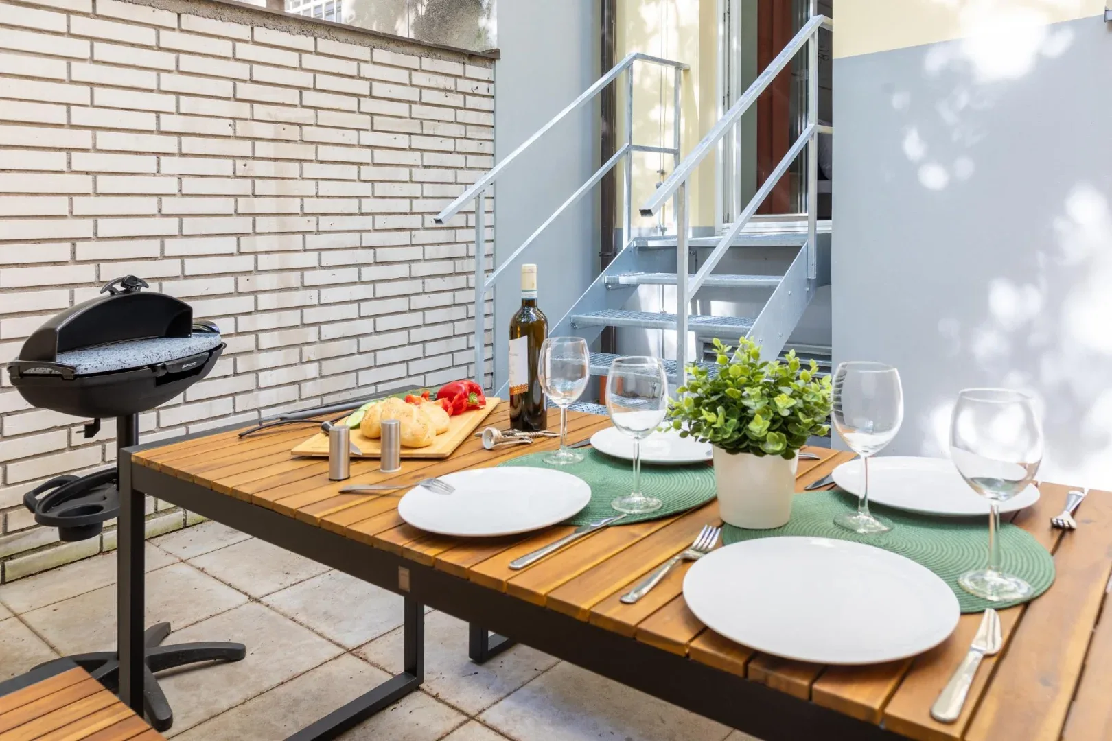 A wooden outdoor dining table is set with plates and glasses beside a barbecue grill and metal stairs.