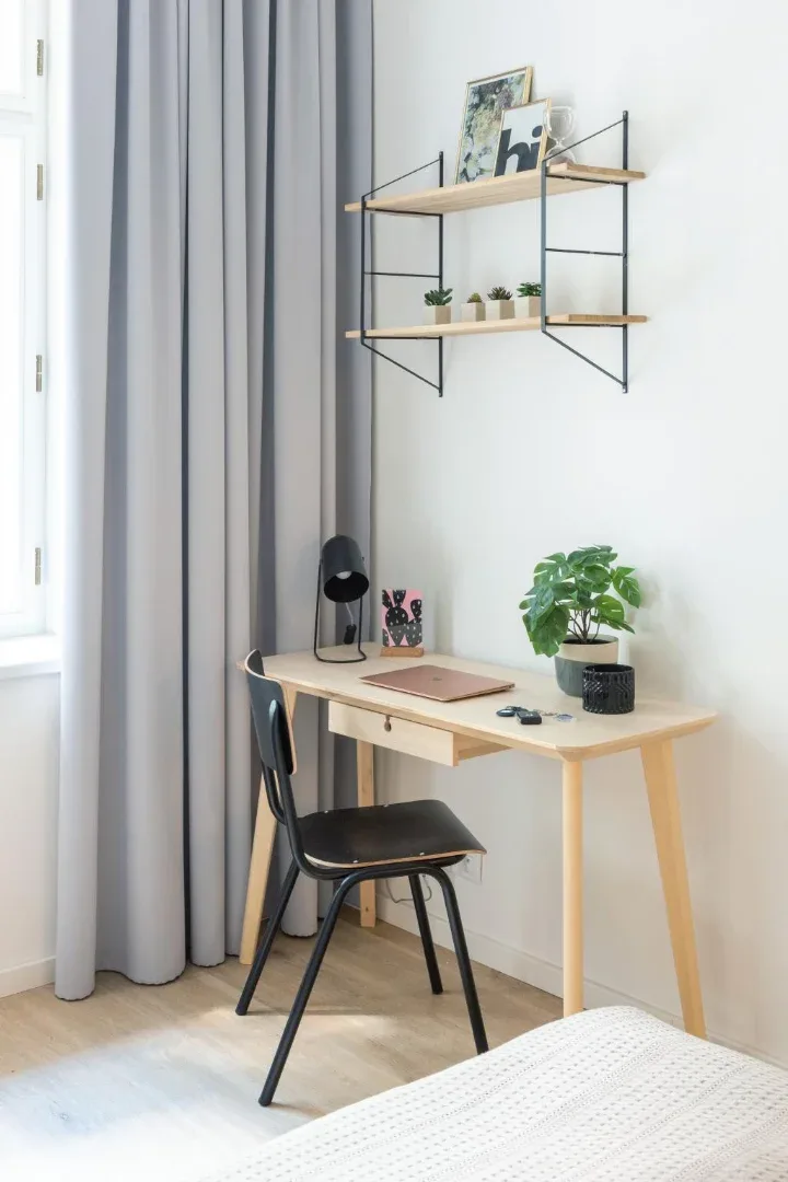 A compact desk with a black chair, lamp, potted plant, and wall shelves sits beside a curtained window.
