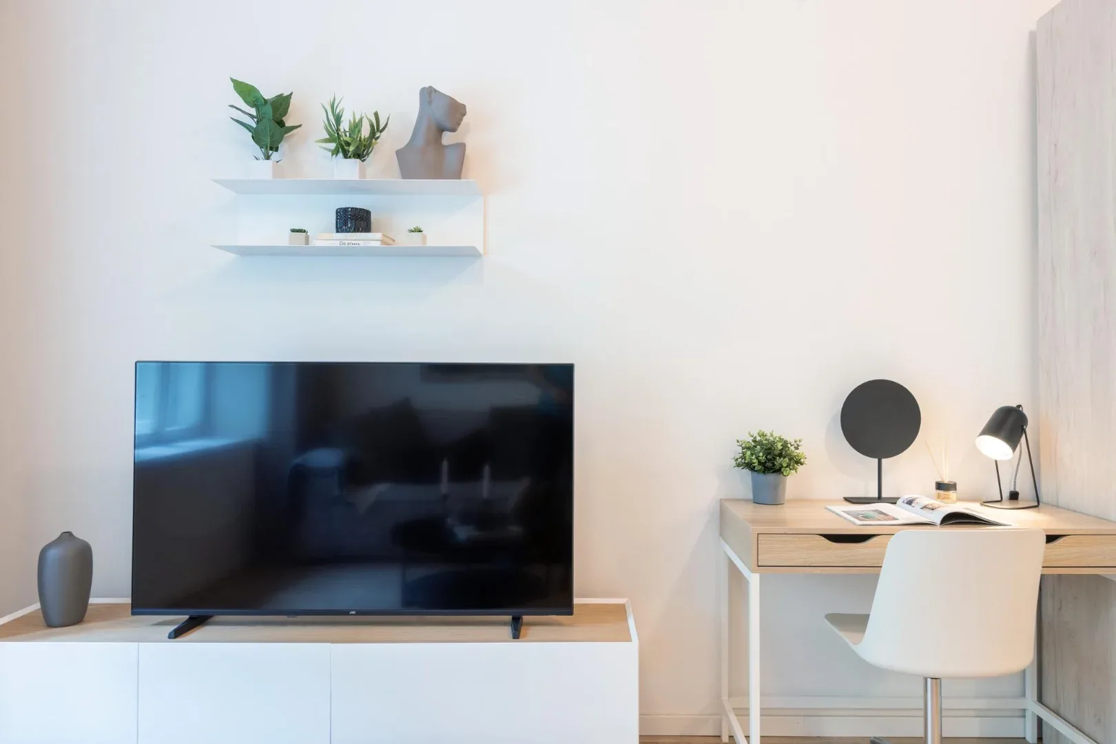 A bright room features a television on a low cabinet and a desk with a white chair and small plants.