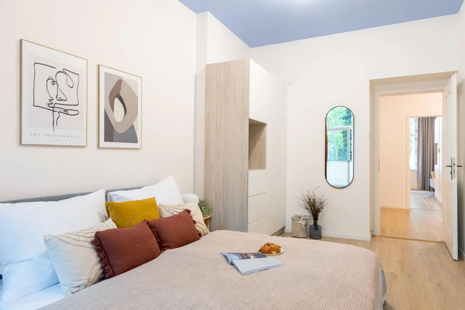 A bright bedroom has a double bed with patterned bedding, framed artwork, and a built-in wooden wardrobe.