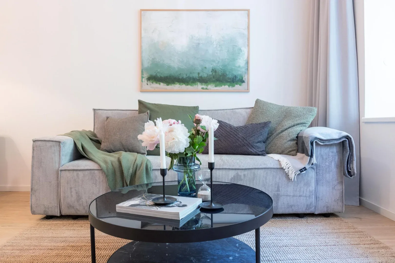 A bright living room features a gray sofa with cushions, a round black coffee table, and flowers.