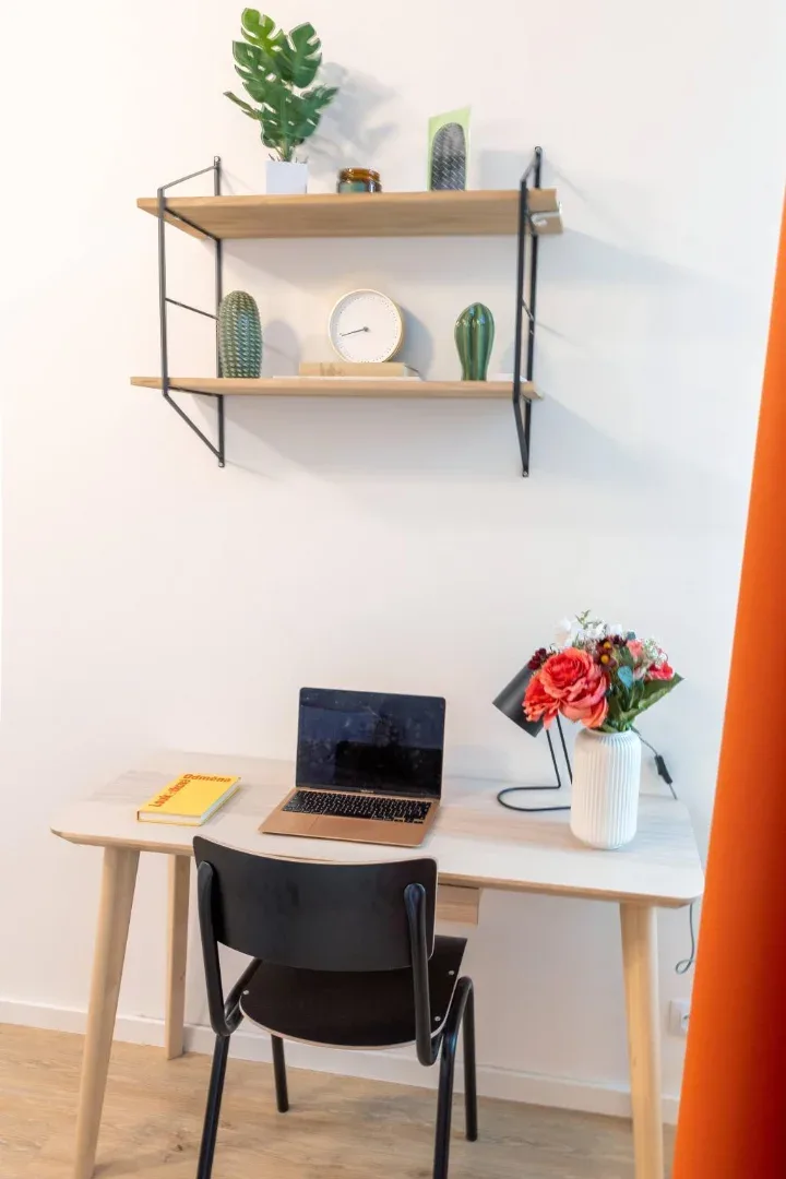 A small desk with a laptop and chair sits beneath wall shelves displaying plants and decorative objects.