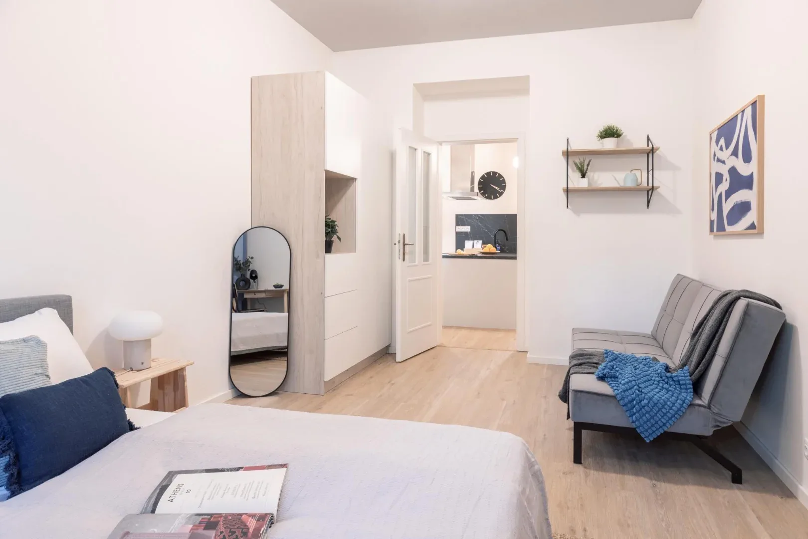 A bright bedroom with a bed, gray sofa, wardrobe, and an open doorway to a small kitchenette.