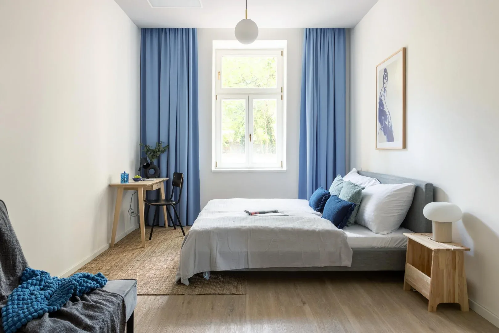 A bright bedroom with a neatly made bed, blue curtains, and a small desk beside the window.