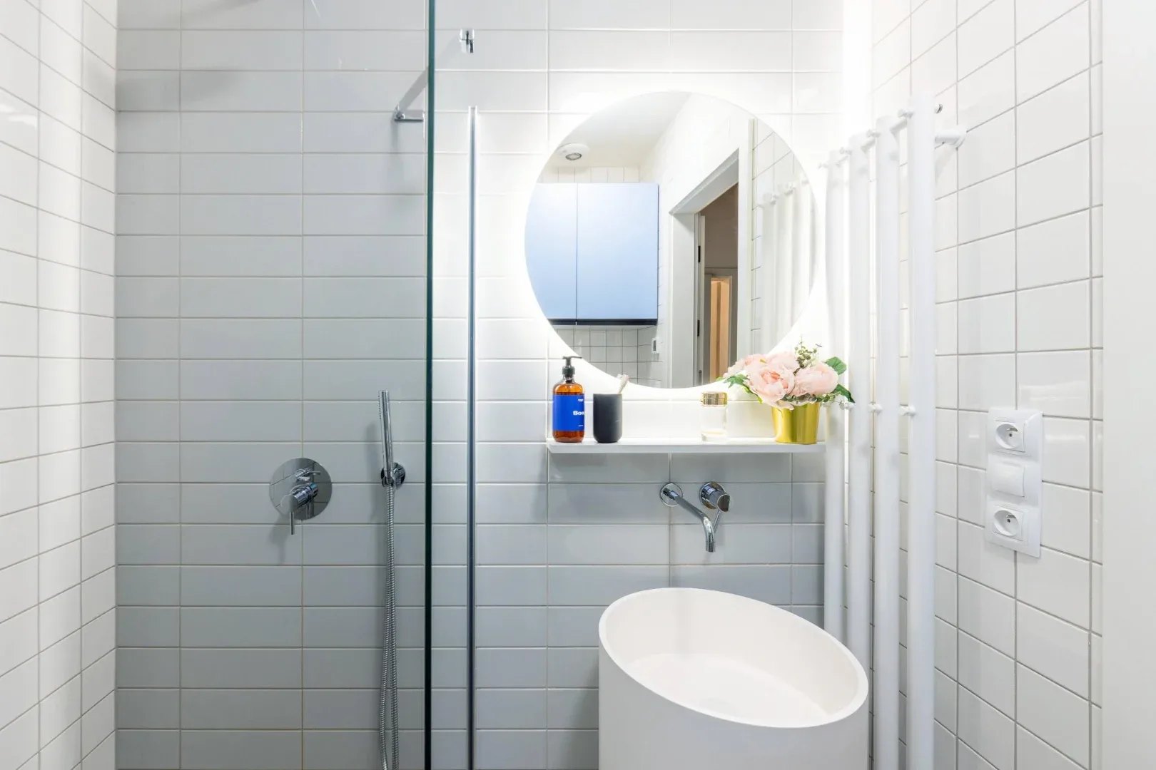 A tiled bathroom features a glass shower, round mirror, wall-mounted faucet, and white vessel sink.