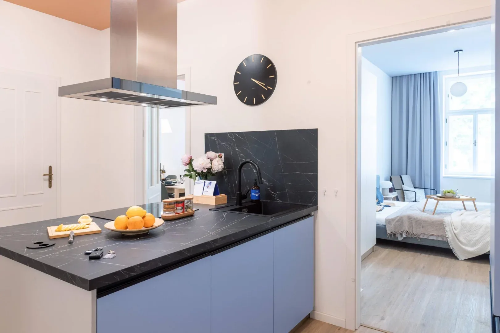 A modern kitchen has a dark stone island, blue cabinets, a stainless-steel range hood, and a round wall clock.